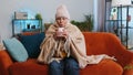 Sick senior grandmother woman wrapped in plaid sit alone shiver from cold on sofa drinking hot tea