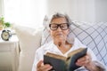 Sick senior woman with glasses reading bible in bed at home or in hospital. Royalty Free Stock Photo