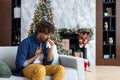 Sick sneezing man with runny nose sitting on sofa at home, hispanic winter day near Christmas tree, cold and allergy in Royalty Free Stock Photo