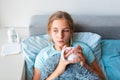 Sick teenage little girl with high fever and headache staying in bed and holding hot cup of tea