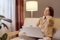 Sick tired woman in knitted jumper using laptop suffering neck pain massaging painful back sitting on sofa in living room in home Royalty Free Stock Photo