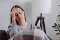 Sick unhappy brunette young woman covered in blanket sitting on couch in living room at home, touching head, suffering from Royalty Free Stock Photo