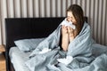 Sick woman froze and caught a cold from an unheated flat covered with warm blanket sit in bed drinking cup with hot beverage or Royalty Free Stock Photo