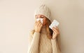 Sick woman sneezing blow nose using tissue and holding pills wearing warm soft knitted clothes, hat and sweater on beige studio Royalty Free Stock Photo