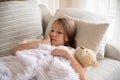 A sick worried little girl covered in blanket is hugging teddy bear.