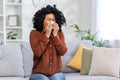 Sick young African-American woman sitting on sofa at home and wiping nose from runny nose with tissue, not feeling well Royalty Free Stock Photo