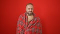 Sick young man suffering from cold, fever and headache wrapped in a blanket over a red isolated background