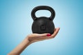 Side closeup of woman`s hand facing up and holding big black 32 kg kettlebell on light blue gradient background. Royalty Free Stock Photo