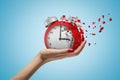 Side closeup of woman`s hand facing up and holding red retro alarm clock that is dissolving into small pieces on light