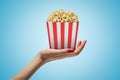 Side closeup of woman`s hand facing up and holding striped popcorn box on light blue gradient background.