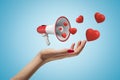 Side closeup of woman`s hand facing up and levitating small megaphone that emits cute red hearts, on light blue