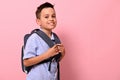 Side portrait of a smiling with toothy smile schoolboy with a school bag on his back on pink background with copy space. Back to
