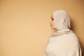 Side portrait of young Arab Muslim woman in strict religious attire and head covered in hijab looking to the side against beige