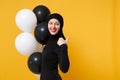 Side profile view Arabian muslim woman in hijab celebrating hold black white air balloons isolated on yellow background