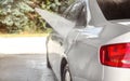 Side of silver car washed in self serve carwash, jet water spraying on metal, blurred sun lit trees background