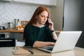 Side view of angry female freelancer talking on mobile phone and using laptop sitting at table in kitchen with modern Royalty Free Stock Photo