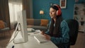 Side view of an Asian woman in red wireless headphones working at a computer. A young woman plays an online game, works