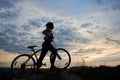 Side view athletic girl in helmet with bicycle on rock under beautiful evening sky with clouds