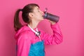 Side view of attractive brunette woman drinking water from bottle, adorable fitness girl posing isolated over pink background, Royalty Free Stock Photo