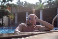 Side view of attractive young bald bearded man adjusting blue sunglasses, standing in swimming pool, enjoying weather. Royalty Free Stock Photo