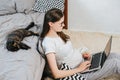 Side view of attractive young pregnant woman in eyeglasses leaning on cozy bed is working on laptop at home near lies cute cat Royalty Free Stock Photo