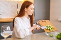 Side view of attractive young redhead woman throwing sliced cucumbers in glass bowl sitting at table in kitchen room. Royalty Free Stock Photo