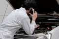 Side view of automotive repairman in uniform talking on phone and examining engine under hood of car at the repair garage.