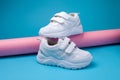 side view balance of two white unisex sneakers on a pink long paper roll on a blue background, one sneaker stands on the