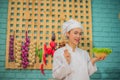 Side view of beautiful asian female professional chef holding wood spatula and a bowl of fresh green vegetables in kitchen. Royalty Free Stock Photo