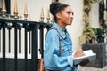 Side view of beautiful casual African American student girl in denim jacket with backpack dreamily writing notes on Royalty Free Stock Photo