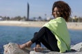 Side view of a beautiful curly afro woman sitting on breakwater rocks laughing while looking camera outdoors Royalty Free Stock Photo