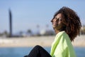 Side view of a beautiful curly afro woman sitting on breakwater rocks laughing while looking camera outdoors Royalty Free Stock Photo