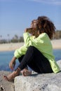 Side view of beautiful curly afro woman sitting on breakwater rocks laughing while resting hand under chin eyes closed outdoors Royalty Free Stock Photo