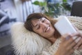 Side view of a beautiful young smiling woman lying on leather couch relaxing while using a mobile phone at home Royalty Free Stock Photo