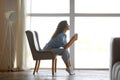 Side view of beautiful young woman drinking hot coffee, sitting on chair near window at home, free space Royalty Free Stock Photo