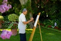 Side view of biracial senior man painting with watercolors on canvas against plants in yard Royalty Free Stock Photo