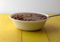 Side view of a bowl filled with baked beans and onions in a white bowl with a spoon in the food atop a yellow tabletop against a