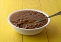 Side view of a bowl filled with baked beans and onions in a white bowl with a spoon in the food atop a yellow tabletop illuminated