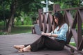 A side view of a brown-haired woman sitting on a wooden bridge in a park using a laptop computer. A young office worker talking on