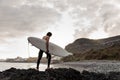 Side view brunette boy walking in the black swimsuit with naked torso with a white surf in his hands on the shore Royalty Free Stock Photo