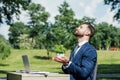 View of businessman with closed eyes holding flowerpot while sitting in park at table with laptop