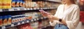 Side view of caucasian woman scans qr code of product using smartphone. Shelves with food in background. Copy space and mock up,