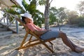 Side view of caucasian young man with hat on face resting on deck chair against trees at beach