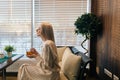 Side view of charming blonde female enjoying cup of hot herbal tea sitting in modern cafe and looking outside through Royalty Free Stock Photo
