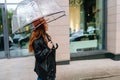 Side view of charming redhead young woman in elegant hat walking on beautiful city street with transparent umbrella Royalty Free Stock Photo