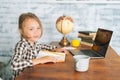 Side view of cheerful cute elementary child school girl doing writing homework sitting at table with laptop and paper Royalty Free Stock Photo