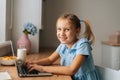 Side view of cheerful elementary child girl using laptop sitting at home table with snack by window, looking at camera. Royalty Free Stock Photo