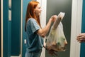 Side view of cheerful redhead female customer receiving groceries bag from unrecognizable delivery man on doorstep at Royalty Free Stock Photo