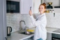 Side view of cheerful redhead young woman opening door of kitchen cabinet at light kitchen room. Royalty Free Stock Photo