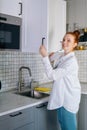 Side view of cheerful redhead young woman opening door of kitchen cabinet at light kitchen room. Royalty Free Stock Photo